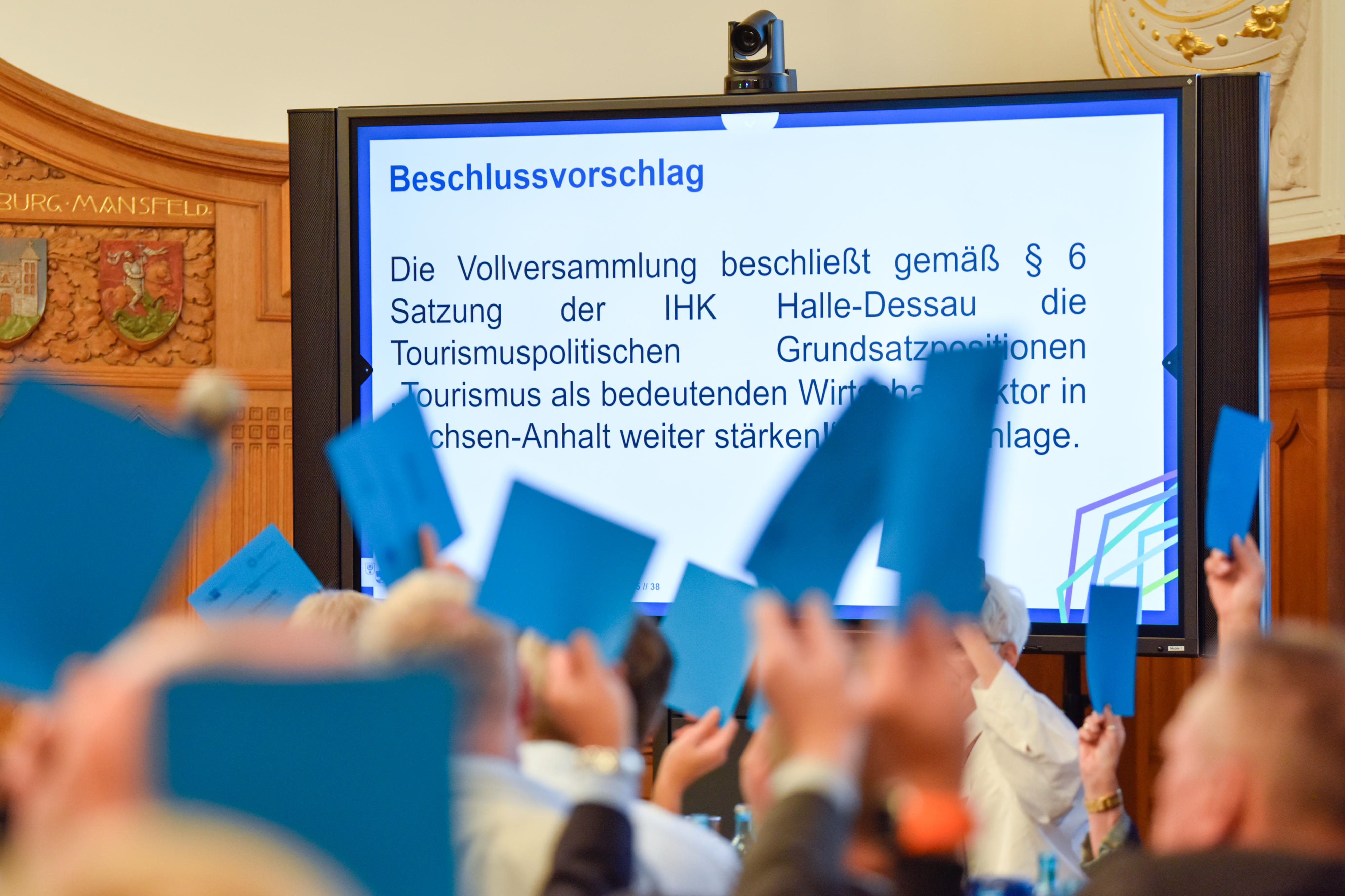 Auf einem Monitor steht Beschlussvorschlag und anwesende Personen halten blaue Abstimmungskarten hoch