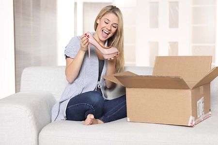 junge Frau freut sich &uuml;ber eine Paketsendung (ge&ouml;ffnetes Paket, Schuh in der Hand)
