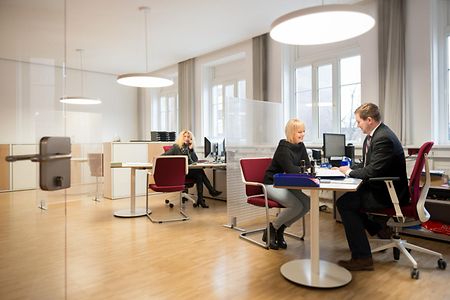 Ein B&uuml;ro mit drei Menschen, in welchem eine Frau von einem Mann beraten wird.