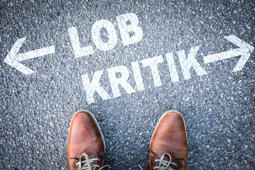 Zwei F&uuml;&szlig;e stehen auf dem Asphalt vor der Schrift "Lob" mit Pfeil nach links und "Kritik" mit Pfeil nach rechts.