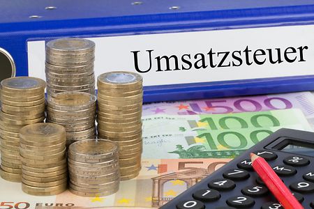 gestapeltes Kleingeld neben einem B&uuml;roordner mit Aufschrift Umsatzsteuer und einem Taschenrechner