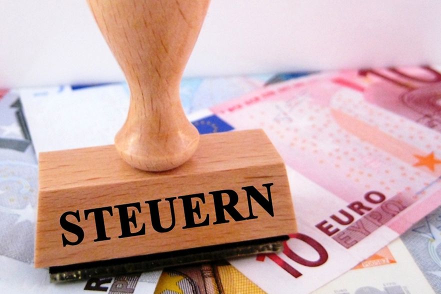 Auf dem Bild ist rechts ein 10 Euro Geldschein und links im Vordergrund ein Stempel mit dem Aufdruck Steuern abgebildet.