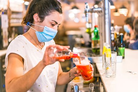 Barkeeperin mit Mundschutz gie&szlig;t Getr&auml;nk in Glas ein.