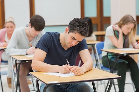 Junge Menschen in einem Klassenraum schreiben eine Pr&uuml;fung. Im Vordergrund ein junger Mann der &uuml;ber seinen Aufgaben sitzt.