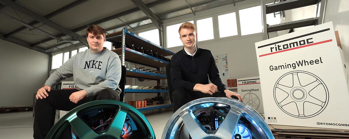 Zwei junge M&auml;nner sitzen in einer Halle vor zwei Gaming Wheels in Felgen Optik.