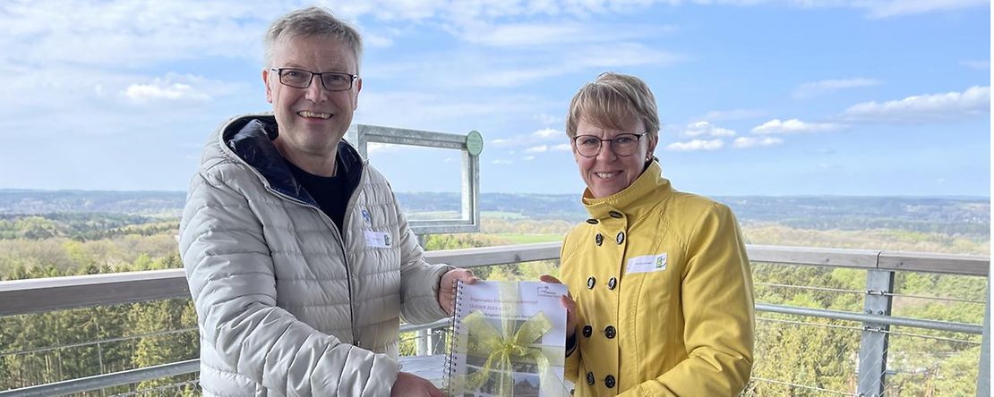 Olaf Muus (l.) und Monika Scherf stehen auf einer Aussichtsplattform und halten eine Urkunde.