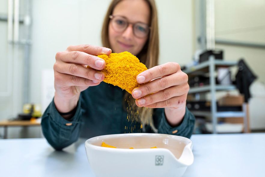 Anne Lamp sitzt an einem Labortisch und br&ouml;selt mit den H&auml;nden gelbes Material in eine wei&szlig;e Schale.