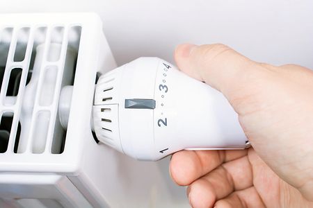 A Male Hand Turns The Radiator Of A White Heater In The Living Room, Saving Energy Concept