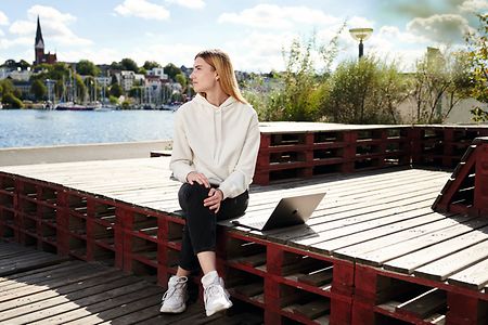 Anna Paramonova sitzt auf einer Holzplattform im Flensburger Hafen mit Laptop neben sich.