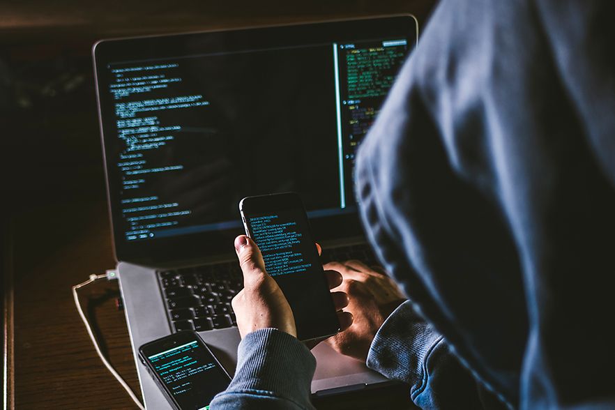 Hacker sitzt vor PC und hat ein Handy in der Hand &ndash; Daten&uuml;bertragung