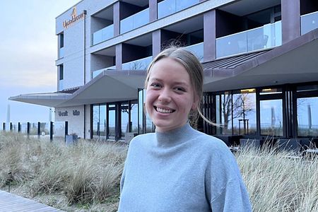 Frau im grauen Pullover steht vor modernem Geb&auml;ude mit Glasfassade.