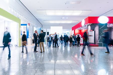 Mehrere Personen stehen in einer Messehalle mit einem roten Messestand im Hintergrund.