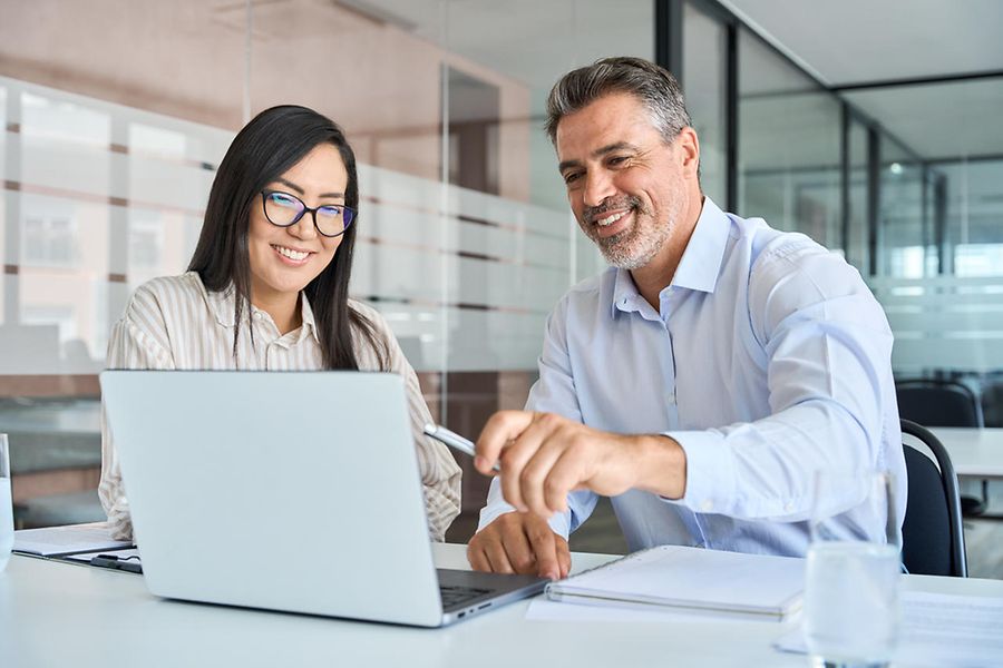 Mature Latin male manager teaching young Asian female worker looking at laptop.