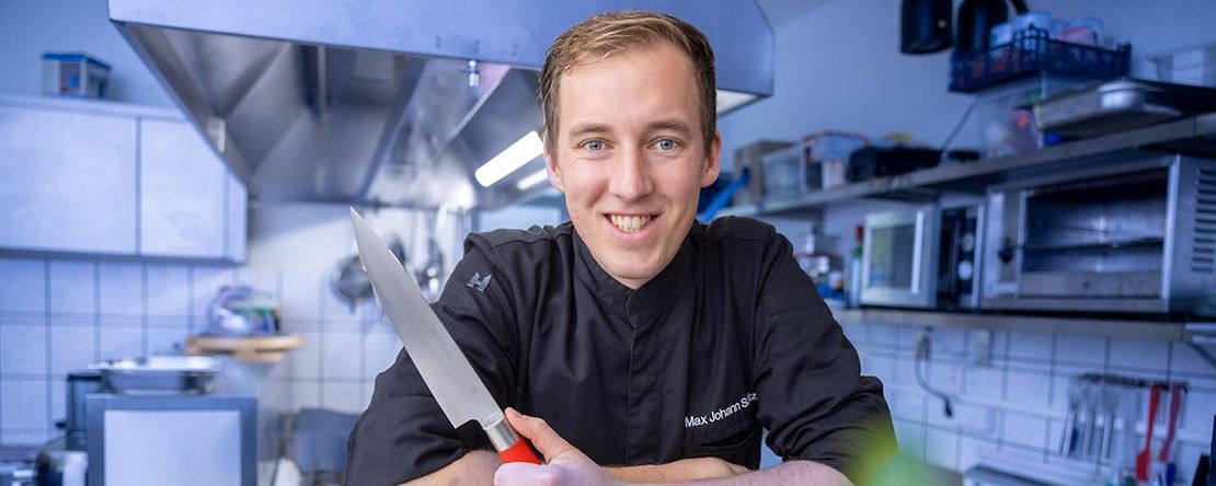 Max Johann Schulze lehnt am K&uuml;chentresen mit einem Messer in der Hand.