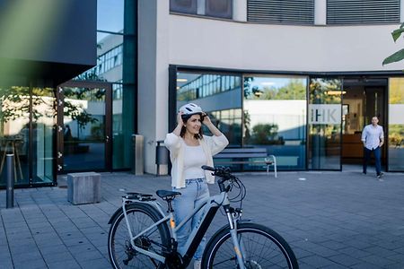 Eine Fahrradfahrerin steht vor dem IHK Geb&auml;ude und zieht gerade Ihren Helm aus.