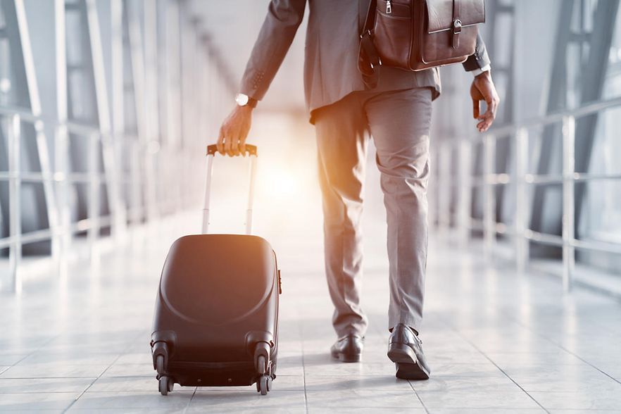 Ein Mann geht durch einen Flur auf einem Flughadfen. Er tr&auml;gt eine braune Ledertasche und zieht einen Koffer hinter sich her.