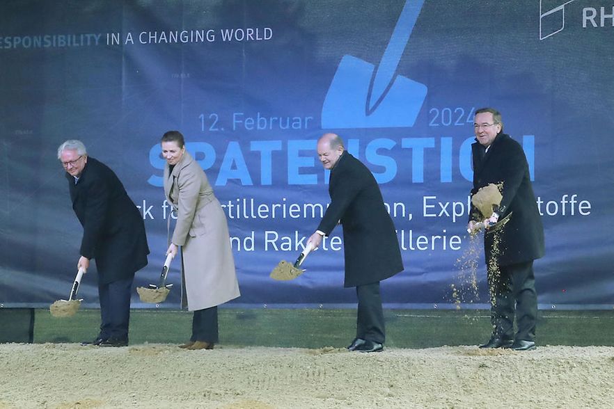 In einem Pavillion aus Plastik-Planen stehen drei M&auml;nner und eine Frau nebeneinander, und werfen mit Spaten Sand auf.