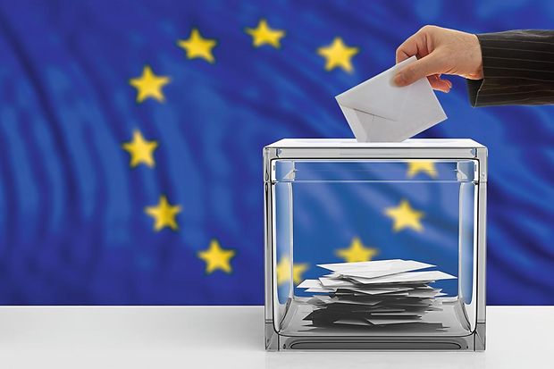 Hand steckt einen Umschlag in eine Wahlurne. Im Hintergrund sieht man die europ&auml;ische Flagge
