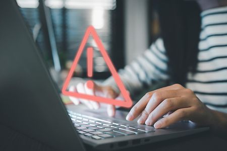 Zwei H&auml;nde tippen auf einer Laptop-Tastatur. Davor ein rotes Achtung-Symbol