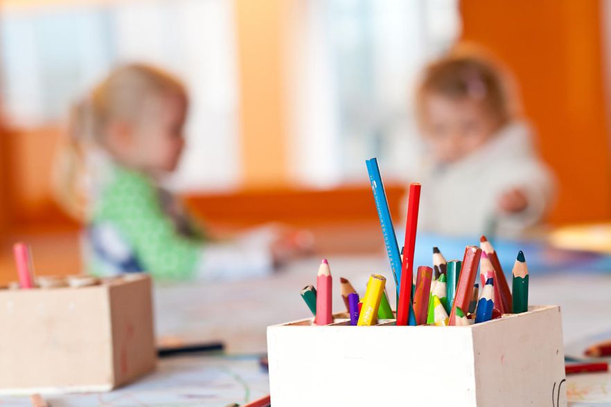 Im Hintergrund sitzen zwei Kinder am Basteltisch einer Kita und malen.