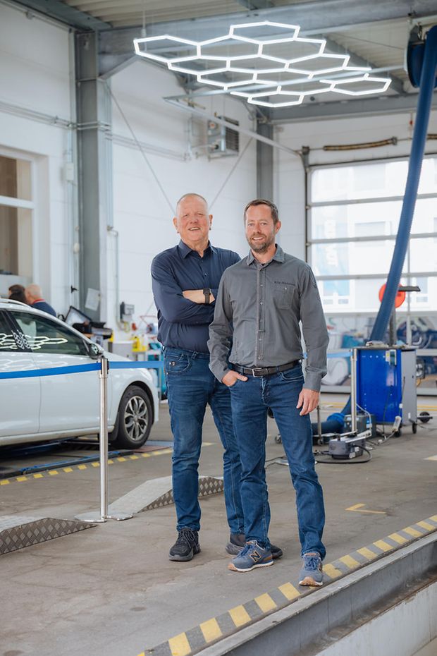 Zwei Männer stehen in einer Autowerkstatt. Sie schauen in die Kamera. Im Hintergrund ein Auto und Werkzeuge.