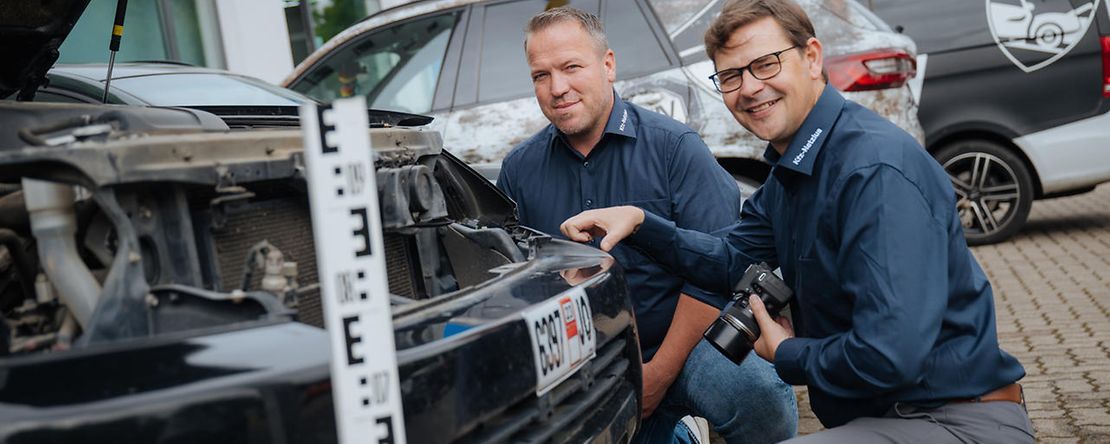 Zwei M&auml;nner in dunkler Kleidung hocken vor einem Auto und schauen in die ge&ouml;ffnete Motorhaube.
