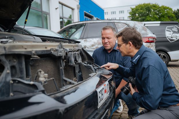 Zwei Männer in dunkler Kleidung hocken vor einem Auto und schauen in die geöffnete Motorhaube.