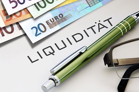 Das Wort "Liquidit&auml;t" steht auf wei&szlig;em Untergrund, daneben liegen ein gr&uuml;ner Stift, eine Brille und Geldscheine.