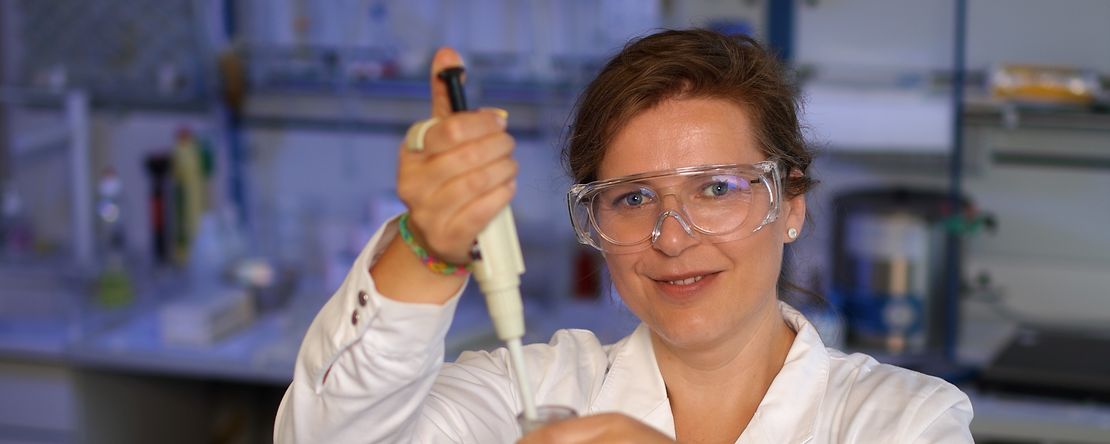 eine Dame mit wei&szlig;em Mantel und Schutzbrille f&uuml;llt etwas in einen Erlenmeyerkolben und l&auml;chelt in die Kamera