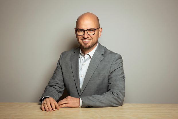 Ein Mann mit Brille und Sakko sitzt an einem Tisch und lächelt in die Kamera.