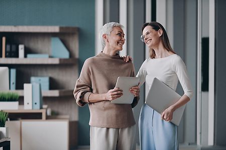 Two beautiful businesswomen holding gadgets and smiling while standing in the office together