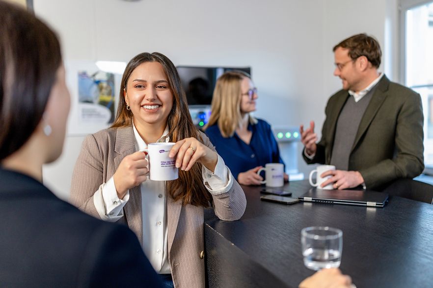 Vier Personen stehen in zwei Gruppen und unterhalten sich an einem Tresen mit Kaffee und Wasser in der Hand.
