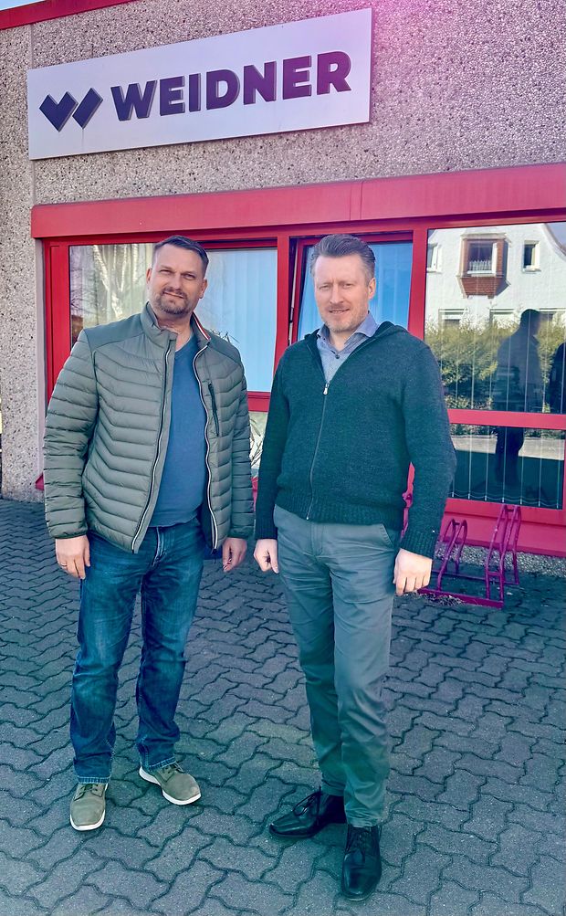 Zwei Männer stehen vor einem Gebäude mit der Aufschrift Weidner.