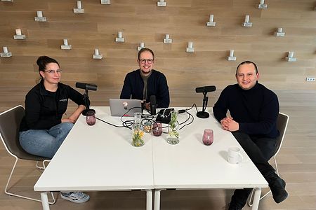 Drei Personen am Tisch mit Mikrofonen und Laptops, bereit f&uuml;r Aufnahme.