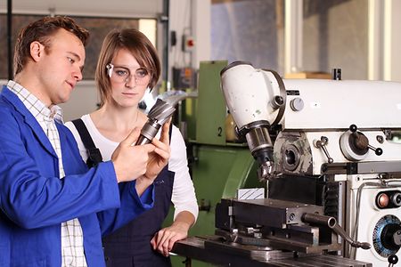 Ein Ausbilder erkl&auml;rt einer Auszubildenden in einer Werkstatt ein Werkzeug an einer Fr&auml;smaschine.