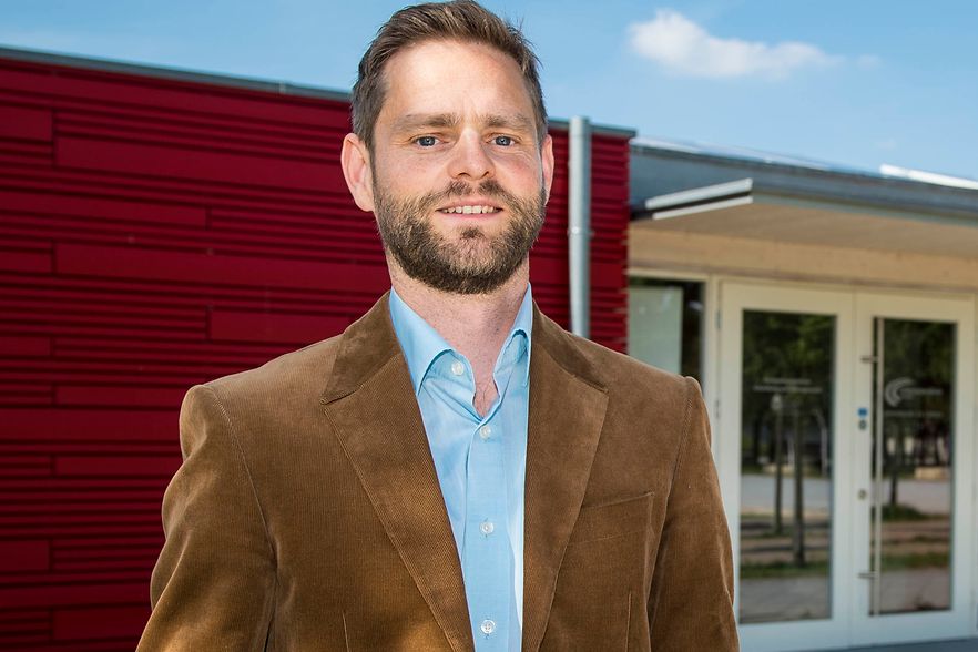 Mann im braunen Sakko vor modernem Geb&auml;ude mit roter Wand.