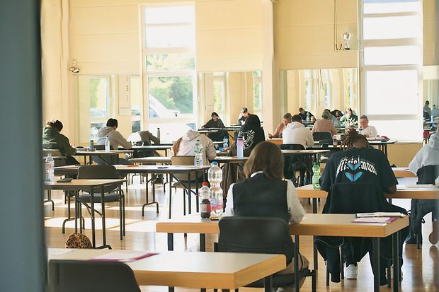 Blick in ein Tanzstudio, in dem Pr&uuml;fungen geschrieben werden. Die Pr&uuml;flinge sitzen an Tischen und arbeiten konzentriert.