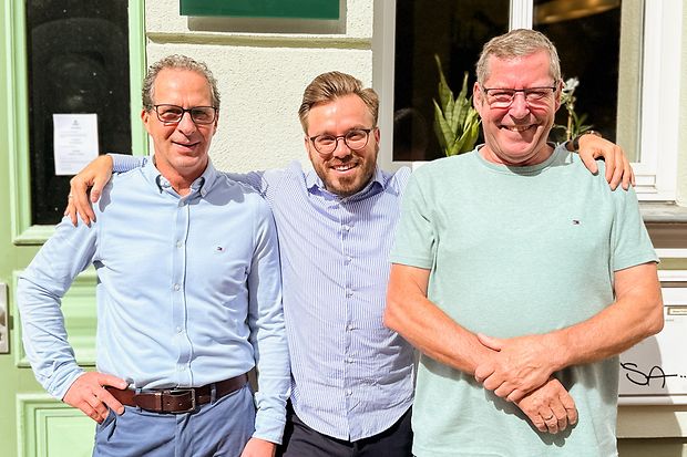 Drei M&auml;nner stehen vor einem Haus und l&auml;cheln in die Kamera.