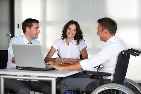 Mann im Rollstuhl sitzt mit zwei Kollegen vor einem Laptop