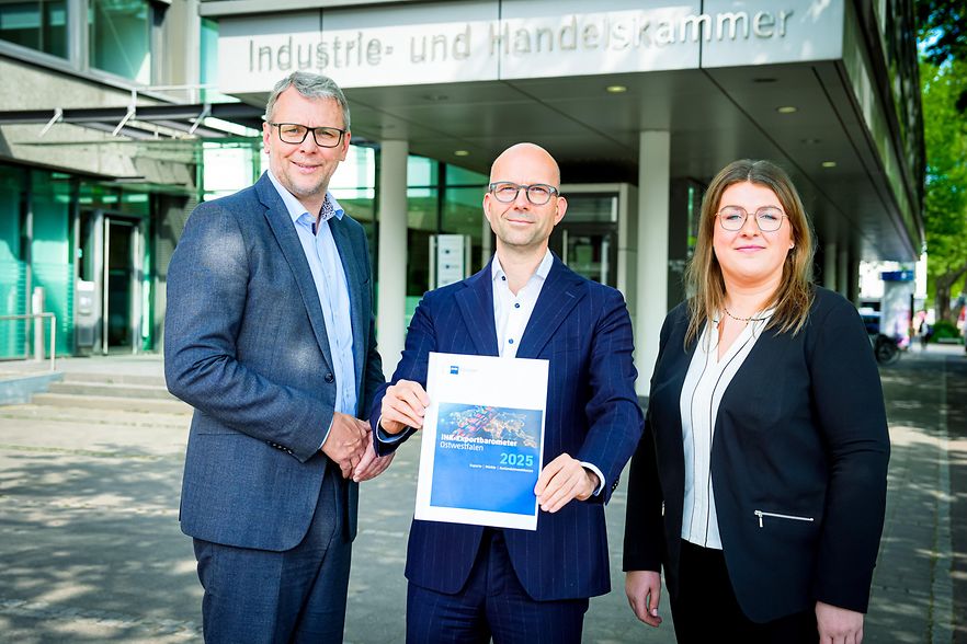 Drei Personen vor der IHK, eine Person h&auml;lt das IHK-Exportbarometer Ostwestfalen 2025.