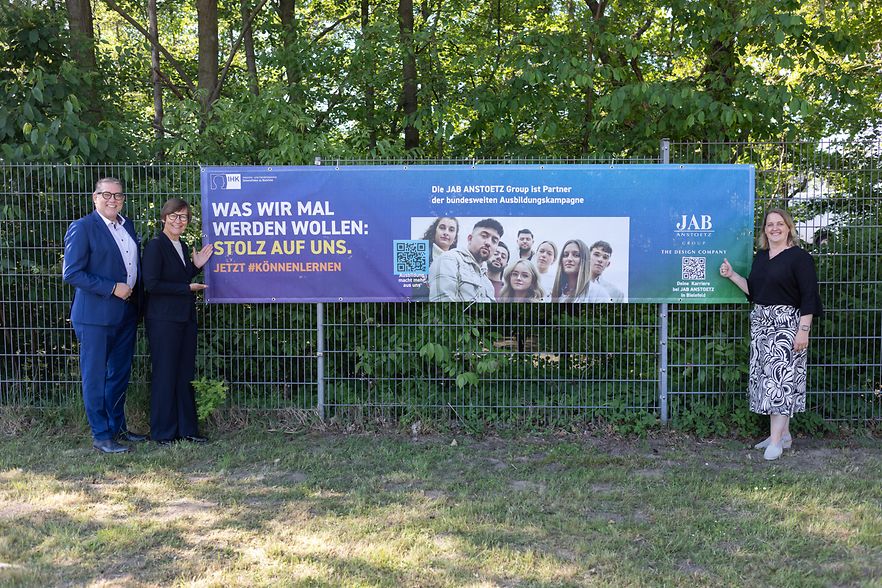 Drei Personen stehen vor einem Zaun mit Banner zur IHK-Ausbildungskampagne bei der JAB Anstoetz Group.