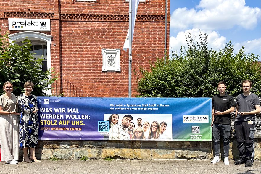 Vier Personen pr&auml;sentieren ein IHK-Ausbildungsbanner vor dem Geb&auml;ude der projekt w Systeme aus Stahl GmbH in Schlo&szlig; Holte.