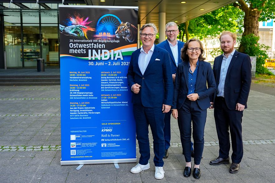 Vier Personen stehen neben einem Roll-Up mit dem Programm der &bdquo;20. IHK-Begegnungstage Ostwestfalen meets India&ldquo; in Bielefeld.