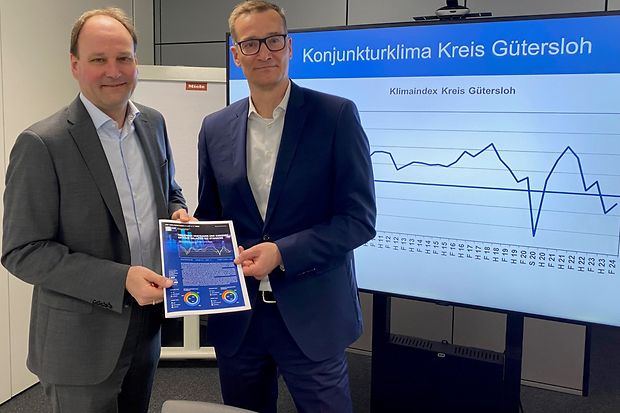 Zwei M&auml;nner im Anzug pr&auml;sentieren Konjunkturbericht vor einem Bildschirm mit Wirtschaftsgrafik Kreis G&uuml;tersloh.