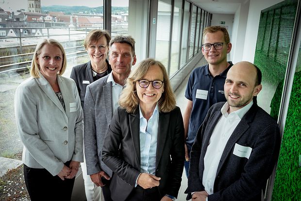 Sechs Personen in Businesskleidung stehen in einem modernen Flur mit Blick &uuml;ber eine Stadt 