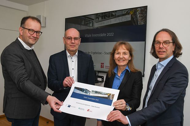 Vier Personen in Businesskleidung stehen vor einem Bildschirm mit der Aufschrift &bdquo;Vitale Innenst&auml;dte 2022&ldquo;.