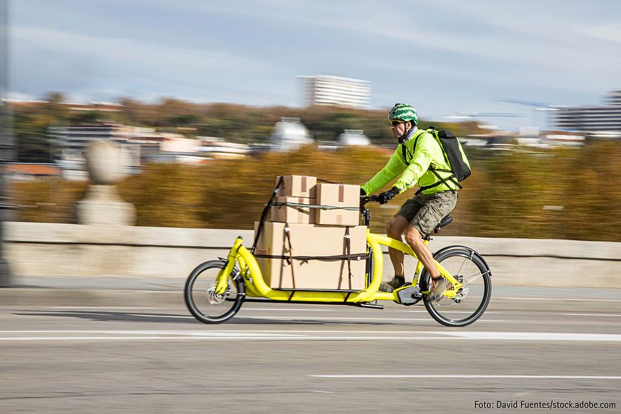 Mann f&auml;hrt mit gelbem Lastenrad, beladen mit Kartons, &uuml;ber eine Stra&szlig;e