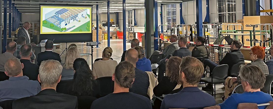 Menschen sitzen in einer Fabrikhalle uns sehen sich eine Pr&auml;sentation &uuml;ber die Firma an.