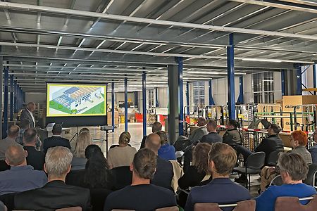 Menschen sitzen in einer Fabrikhalle uns sehen sich eine Pr&auml;sentation &uuml;ber die Firma an.