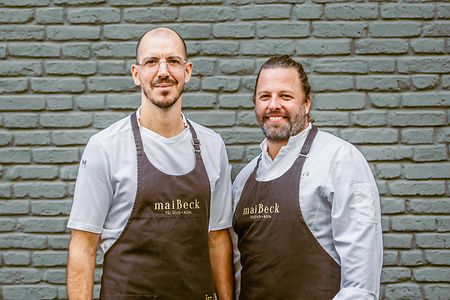 Jan Maier und Tobias Becker tragen Sch&uuml;rzen und stehen vor einer Wand.